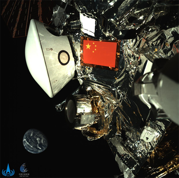 Image publiée par l'Administration spatiale nationale chinoise le 1er octobre 2025 montrant une vue de la sonde Tianwen-2 aux côtés de la Terre, capturée par la sonde lors de son voyage dans l'espace lointain. Cette nouvelle image, acquise par une caméra de surveillance montée sur le bras robotisé de la sonde, met en valeur le drapeau rouge à cinq étoiles de la Chine et la capsule de retour blanche sur fond de Terre bleue lointaine. (Photo fournie par l'Administration spatiale nationale chinoise)